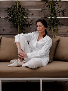 Female model sitting on outdoor furniture in front of a wood wall and bamboo wearing white Jil Sander outfit with Maison Margiela sneakers