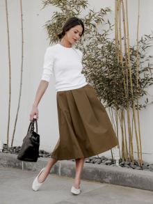 Female model standing in front of bamboo wall wearing white Alexandra Golovanoff sweater, olive pleated Aspesi skirt, white The Row ballet flat, while swinging a brown and black leather Henry Beguelin handbag