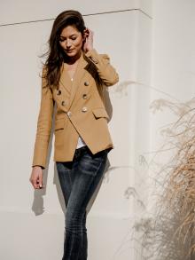 Female model standing against white wall next to fountain grass wearing camel colored Veronica Beard jacket and R13 jeans