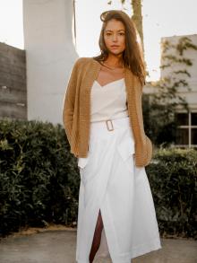 Female model standing in front of bamboo and white architecture at sunset wearing white Vince skirt, camel White+Warren cardigan, and off white silk L’Agence tank