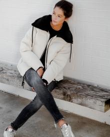 Model sitting on wooden bench wearing white Varley Puffer Jacket, black 6397 Sweater R13, faded blue Skinny Jean, and white Golden Goose Sneaker