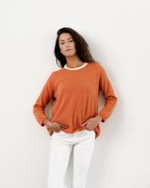 Model standing against white wall wearing orange Bruno Manetti Sweater and white R13 Jean